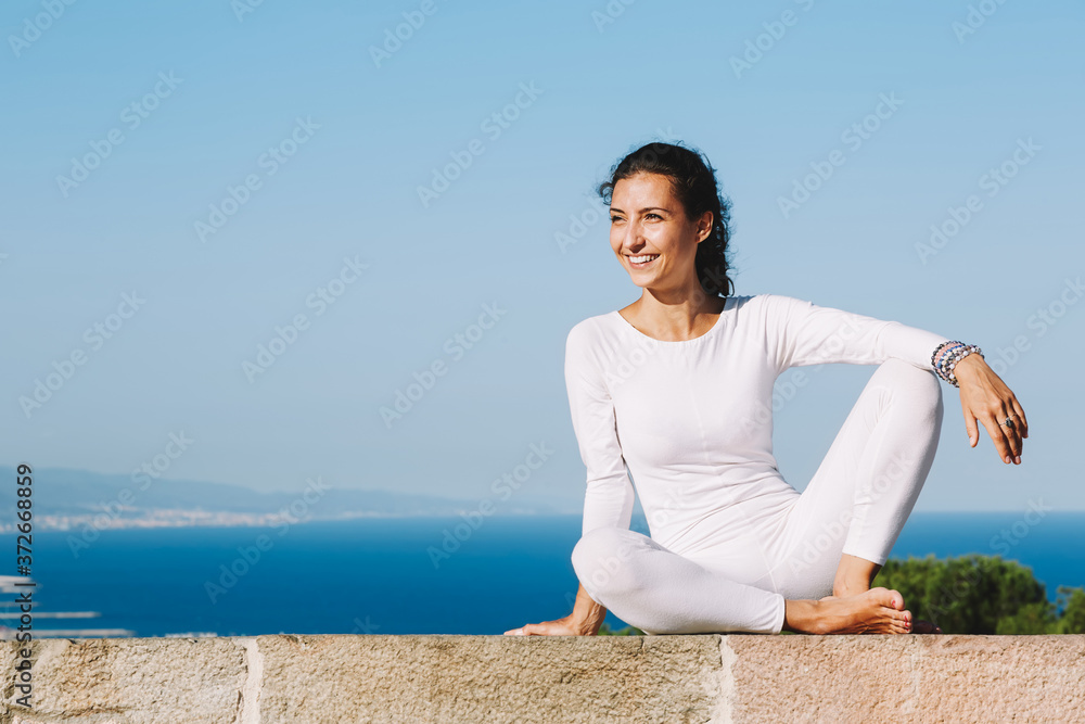 Yoga on high altitude with big city on background, smiling woman seated in yoga pose on amazing city background, woman meditating yoga and enjoying sunny evening, woman makes yoga on mountain hill