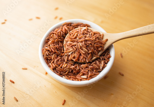 Closeup brown rice or red rice with wooden spoon and white ceramic bowl on wood table top. Healthy organic food concept.