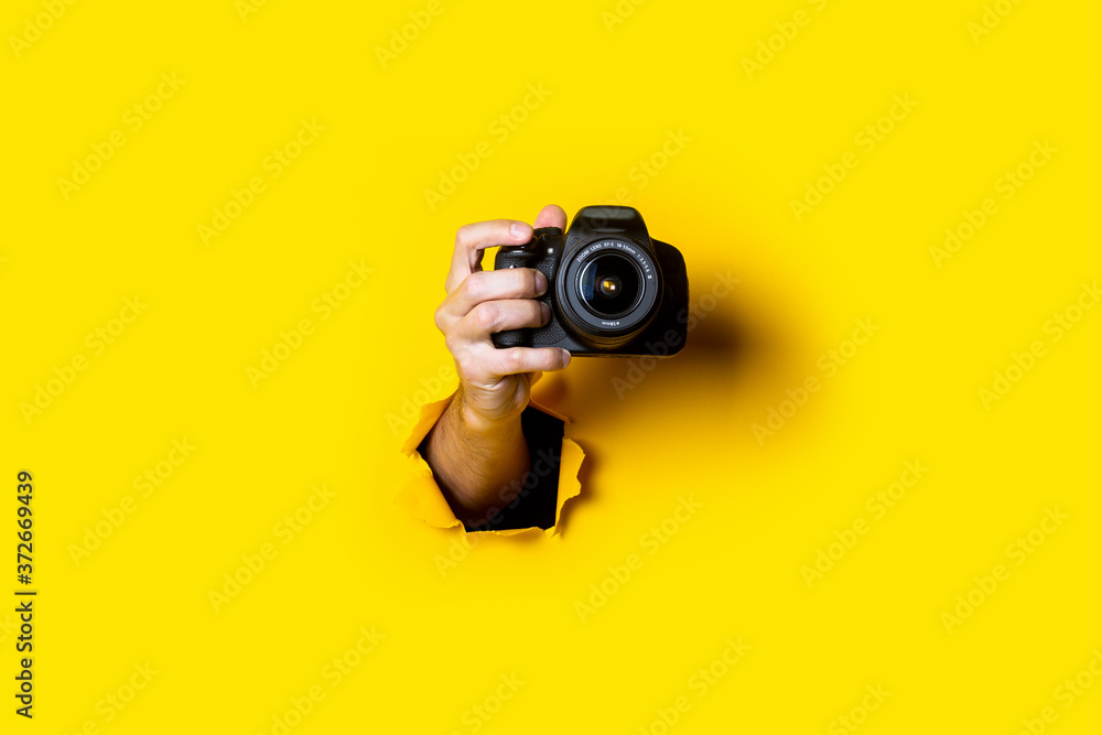 Man's hand holding a camera on a bright yellow background. - obrazy, fototapety, plakaty 