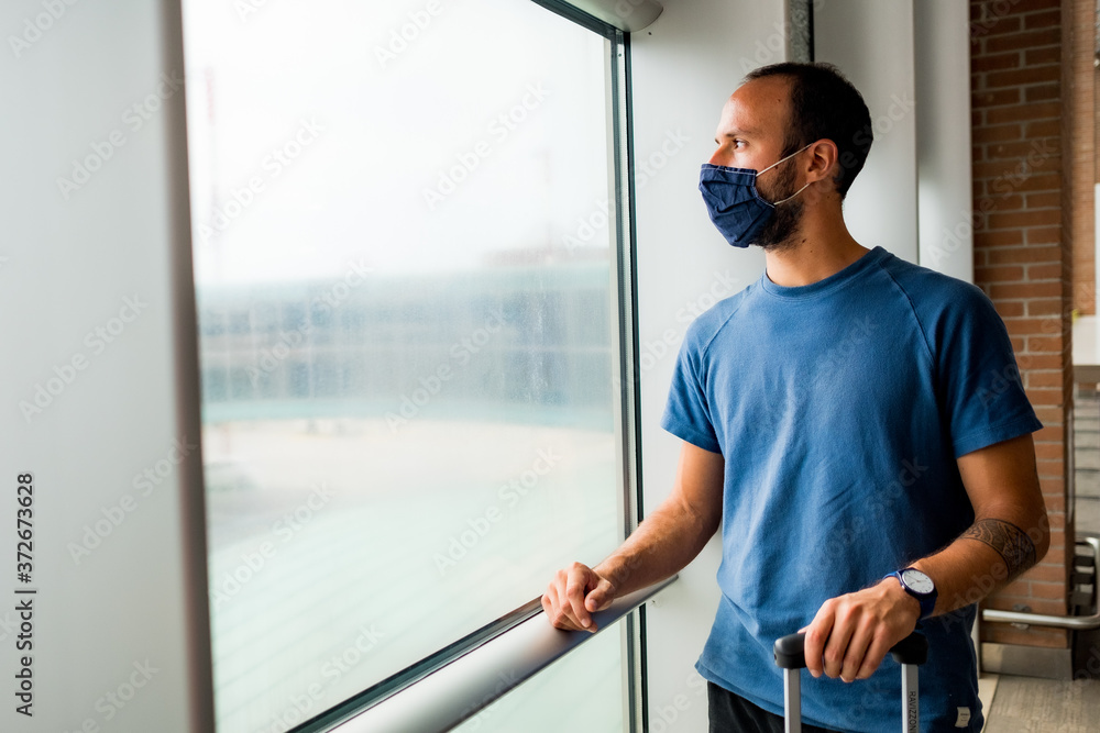 Fototapeta premium young man wearing protective face mask, traveling in the time of covid19 pandemic inside an airport, looking out of window with worried and upset expression. corona virus and travel industry