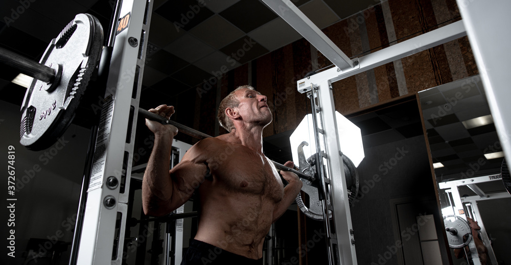 Naklejka premium Muscular adult brutal man doing barbell squats in the gym. Portrait of caucasian authentic bodybuilder doing workout exercises