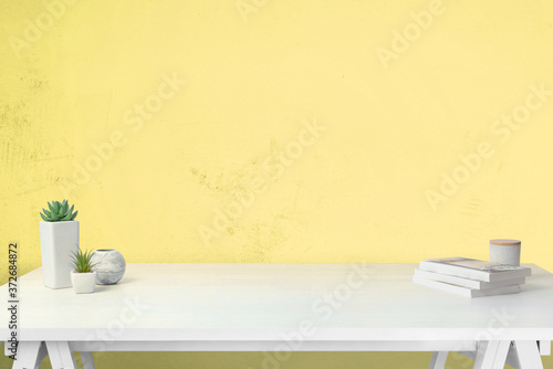 Work desk with free space for product promotion. Yellow wall in background. Books and plants on the table. Front view