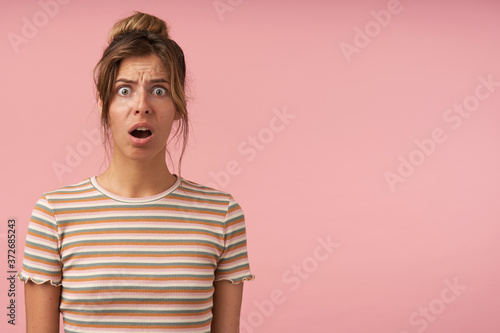 Indoor photo of young bewildered brown haired lady with natural makeup frowning her eyebrows while looking perplexedly at camera and keeping mouth opened, isolated over pink background