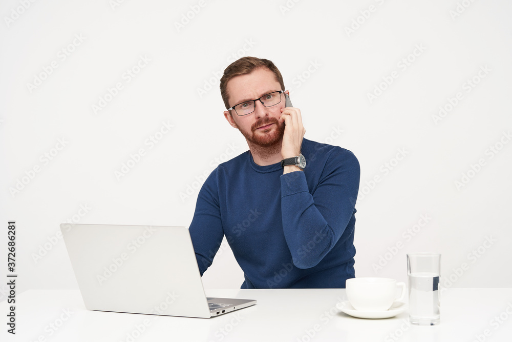 Bewildered young pretty bearded guy dressed in casual clothes having phone conversation and frowning eyebrows while looking confusedly ar camera, isolated over white background Bewildered young pretty bearded guy dressed in casual clothes having phone conversation and frowning eyebrows while looking confusedly ar camera, isolated over white background
