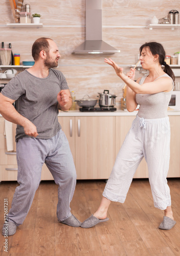 Positive cheerful crazy couple singing and dancing while having breakfast in kitchen wearing pajamas. Carefree wife and husband laughing having fun funny enjoying life authentic married people