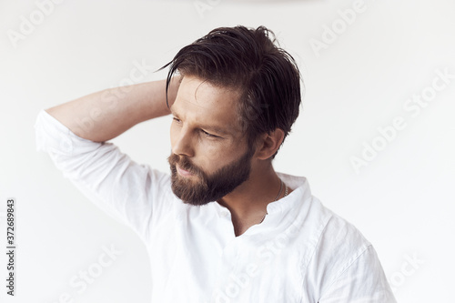 high key portrait photo on white cyclorama of a handsome bearded man with brown hair and eyes, he is wearing a white linen shirt, he holds his hand behind his head and looks away