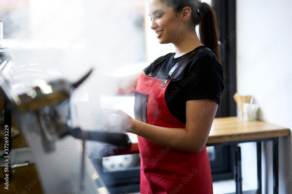 Cheerful waitress in uniform standing at bar enjoying making coffee on machine serving order in good mood.Beautiful female owner of retail business work as barista steaming milk for beverages Cheerful waitress in uniform standing at bar enjoying making coffee on machine serving order in good mood.Beautiful female owner of retail business work as barista steaming milk for beverages