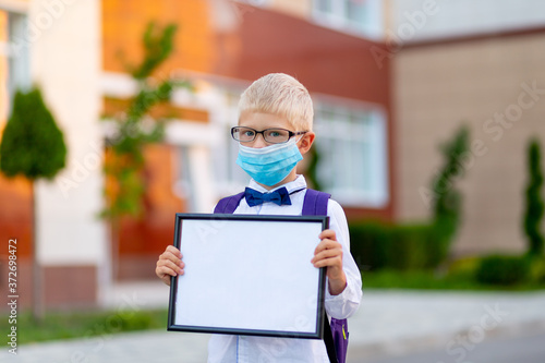 a blond schoolboy with glasses and a protective mask stands at the school and holds a sign with a white sheet. Day of knowledge. Space for text photo