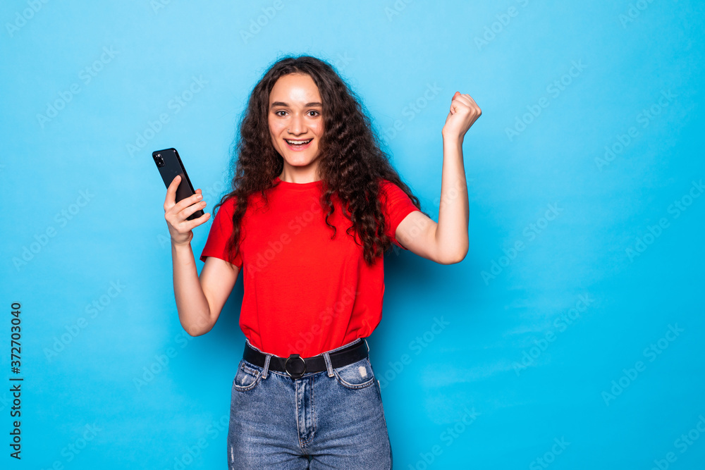 Portrait of a happy beautiful girl looking at mobile phone and celebrating isolated over blue background Portrait of a happy beautiful girl looking at mobile phone and celebrating isolated over blue background