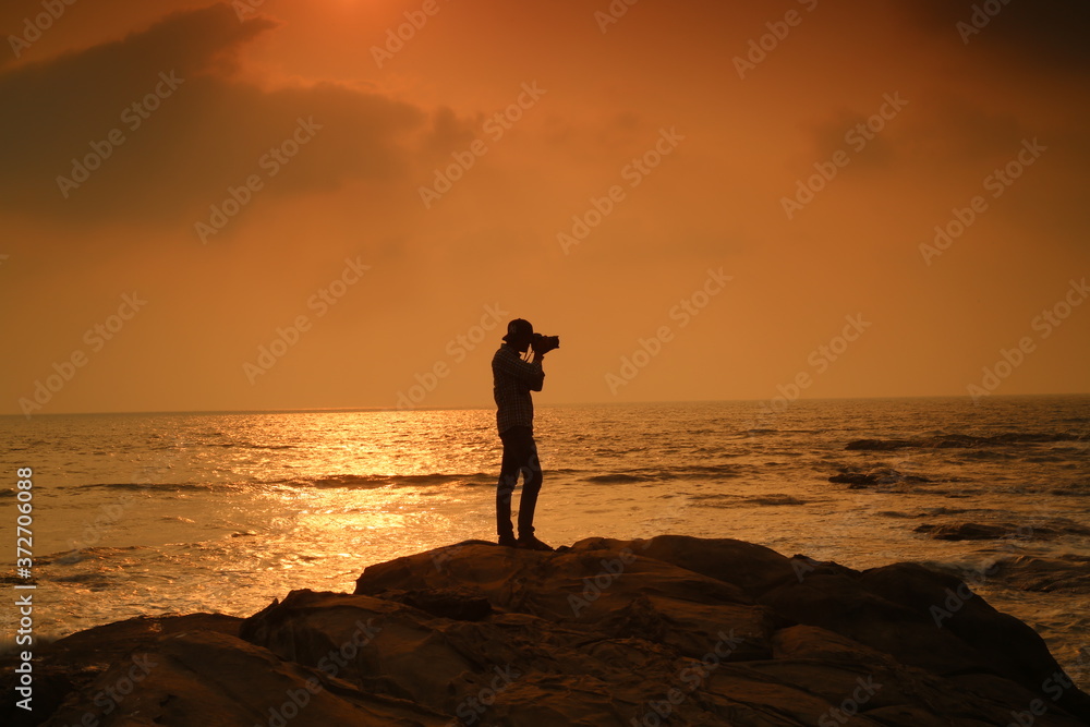 A tourist stand on rock and sea water facing sunset and clicking picture background wallpaper. 