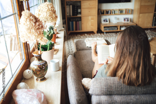 Rear view of brunette young woman reading book on sofa in living room at home