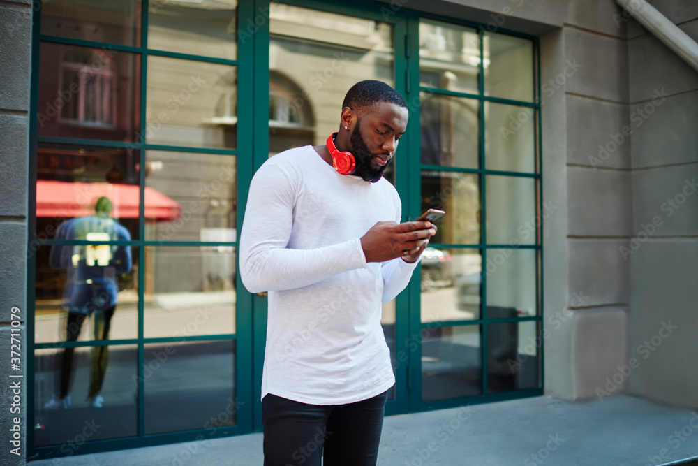 Заголовок: Positive afro american guy with headphones searching favorite songs for downloading on mobile walking on street, cheerful dark skinned male hipster share photos in social networks chatting 