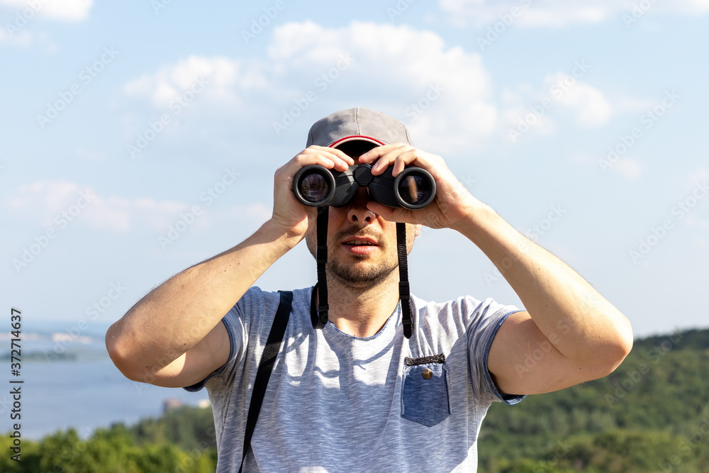 A man looking at camera through binoculars against a scenic view of a wide river on a hill on a sunny day A man looking at camera through binoculars against a scenic view of a wide river on a hill on a sunny day