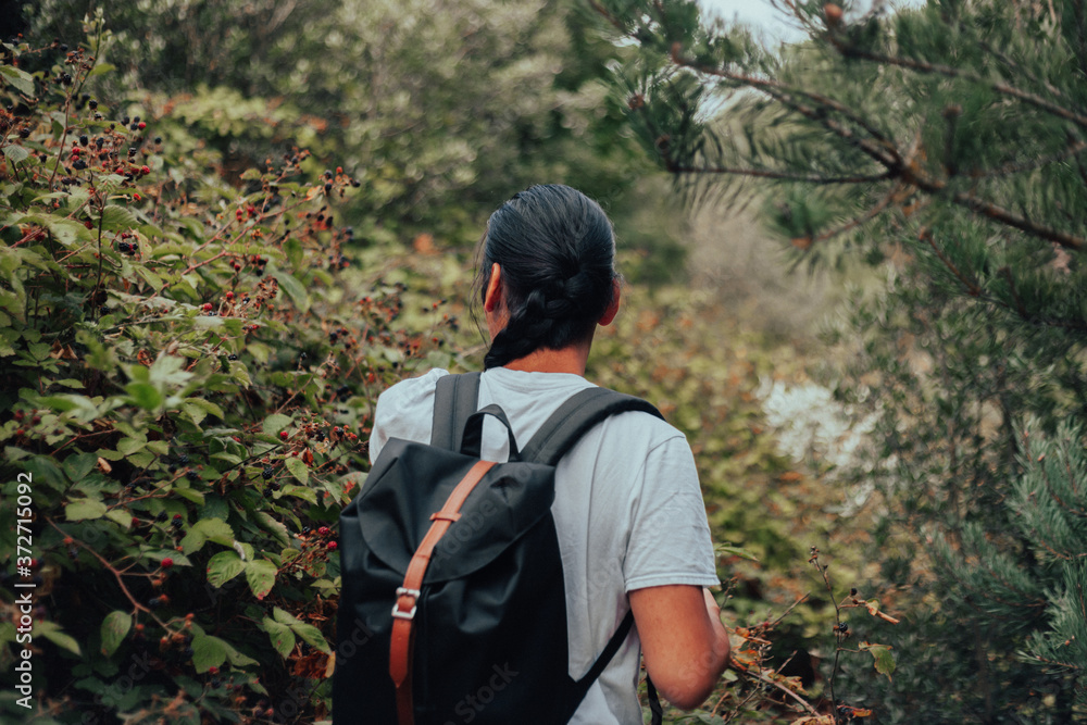 A young tanned man in a light grey T-Shirt, with long black hair and a backpack standing in a blackberry bramble bushes with his back to the camera