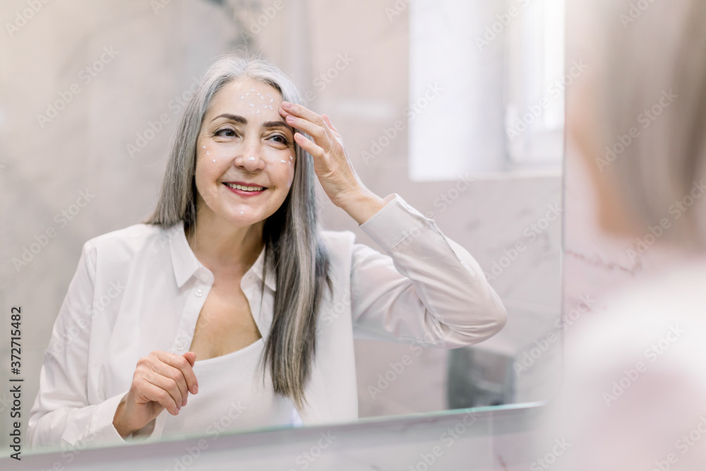 Pretty senior woman with long gray hair, wearing white shirt, looking at her face in the bathroom mirror, and applying anti-wrinkle cream or cosmetic lotion. Skin care, antiaging treatment Pretty senior woman with long gray hair, wearing white shirt, looking at her face in the bathroom mirror, and applying anti-wrinkle cream or cosmetic lotion. Skin care, antiaging treatment