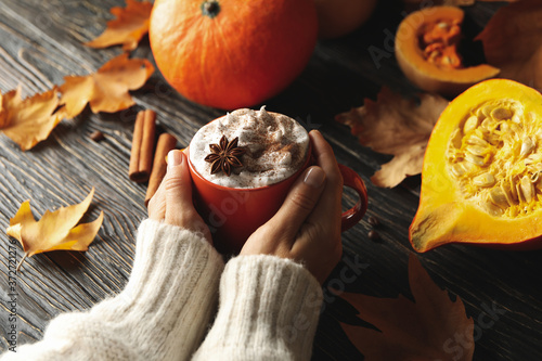 Female hands hold cup of pumpkin latte on wooden background with pumpkin, cinnamon and leaves