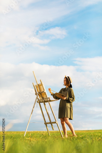Young woman artist paints a picture outdoors on a warm summer evening. Model posing in dress barefoot