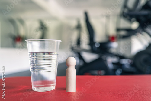 Wooden people man standing near big disposable plastic glass of still water in fitness gym opposite the sport simulators. Resting time. Health concept.