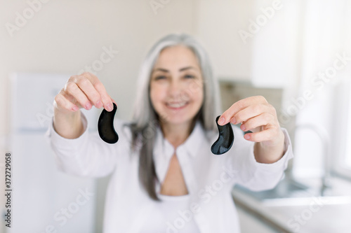 Blurred portrait of happy retired woman with gray hair holding black antiwrinkle eye patches, showing it at camera, while posing in light cozy kitchen at home. Focus on hands with patches