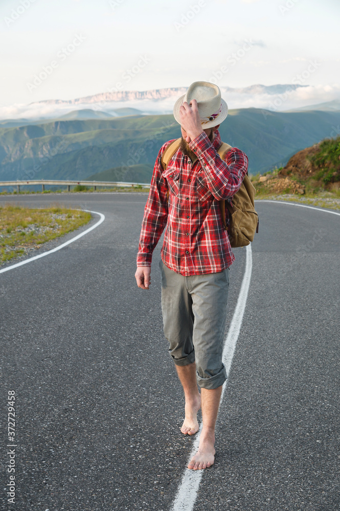 Φωτογραφία A young bearded hipster in a shirt hat and barefoot walks along a country road in the mountains