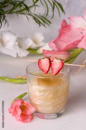 A drink with a shiny shimmer decorated with strawberry wedges next to flowers
