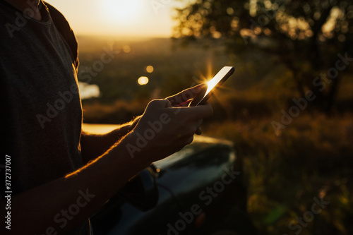 Cropped shot of a young man using smart phone. Man's hands using smart phone at sunset. Cropped shot view of hipster guy reading text message on cell telephone.