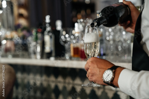waiter pouring champagne into grocers from a bottle against the background of a bar. Alcoholic luxury party. © galaganov