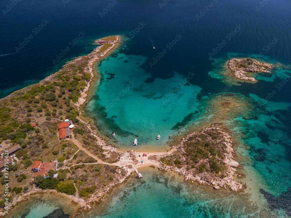 Mediterranean Greek landscape coastal drone shot with moored leisure boats. Aerial day top view of Sithonia Chalkidiki peninsula above small island shoreline with green plantation & clear calm sea.