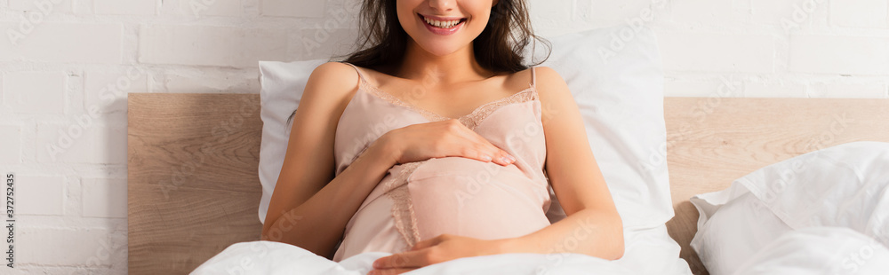 panoramic crop of pregnant woman in silk nightie touching belly in bedroom