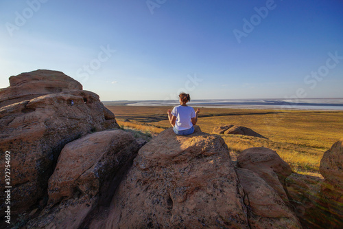 a young woman is sitting on a Large Stone looking into the distance on the steppe on the lake and meditating, view from the back © Ekaterina