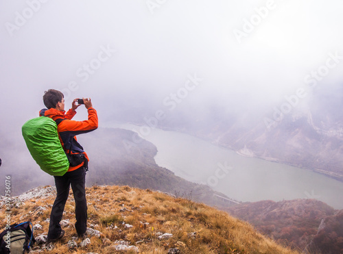 Man hiker on the mountain peak taking photo of Danube river landscape on foggy autumn day