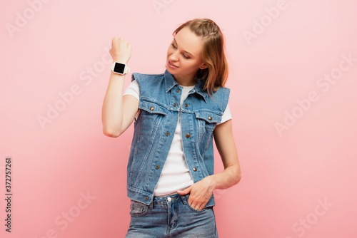 young woman in denim vest showing smartwatch on wrist while standing with hand in pocket isolated on pink
