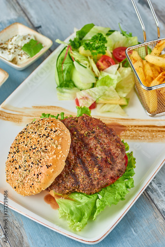 Delicious grilled burger on white plate on wooden table. with sauces, french fries and salads.