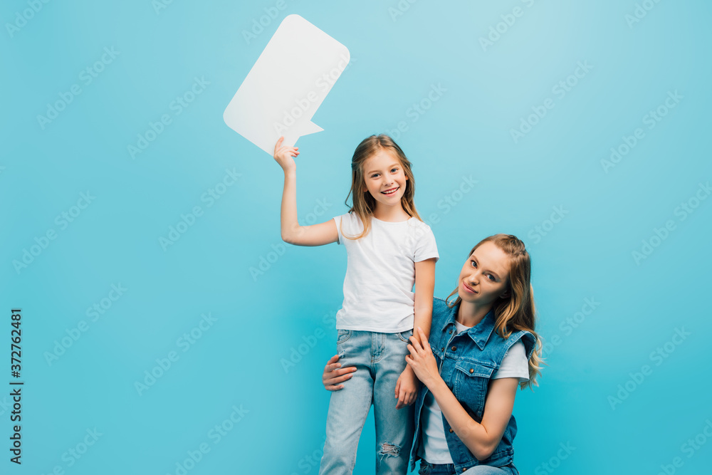 mother squatting near child wearing white t-shirt and jeans and holding speech bubble isolated on blue mother squatting near child wearing white t-shirt and jeans and holding speech bubble isolated on blue