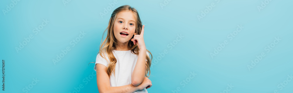 horizontal image of excited girl in white t-shirt showing idea sign while looking at camera isolated on blue