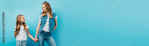 horizontal image of young woman in denim clothes holding hands with daughter isolated on blue