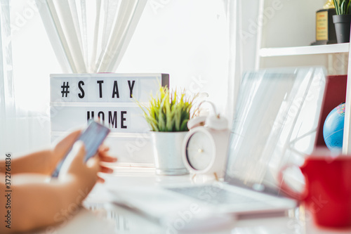 lightbox with text hashtag #STAYHOME glowing in lightand blurred woman working at home. Office worker on quarantine. Home working to avoid virus disease. Freelancer or remote worker concept. photo