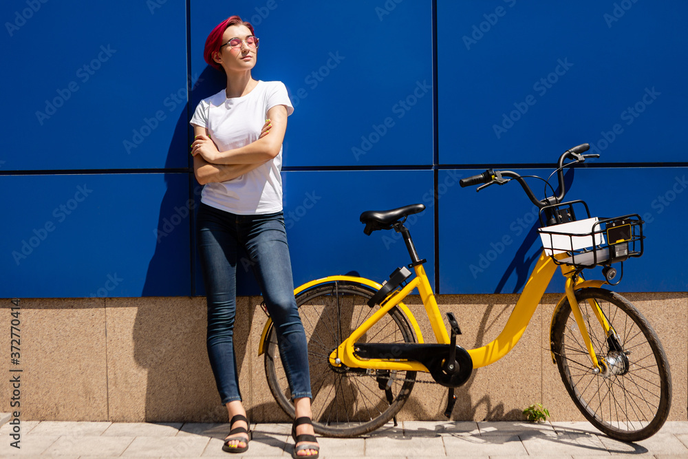 Obraz premium Happy young woman with pink hair walks around the city on a bike in the summer. Eco-friendly mode of transport