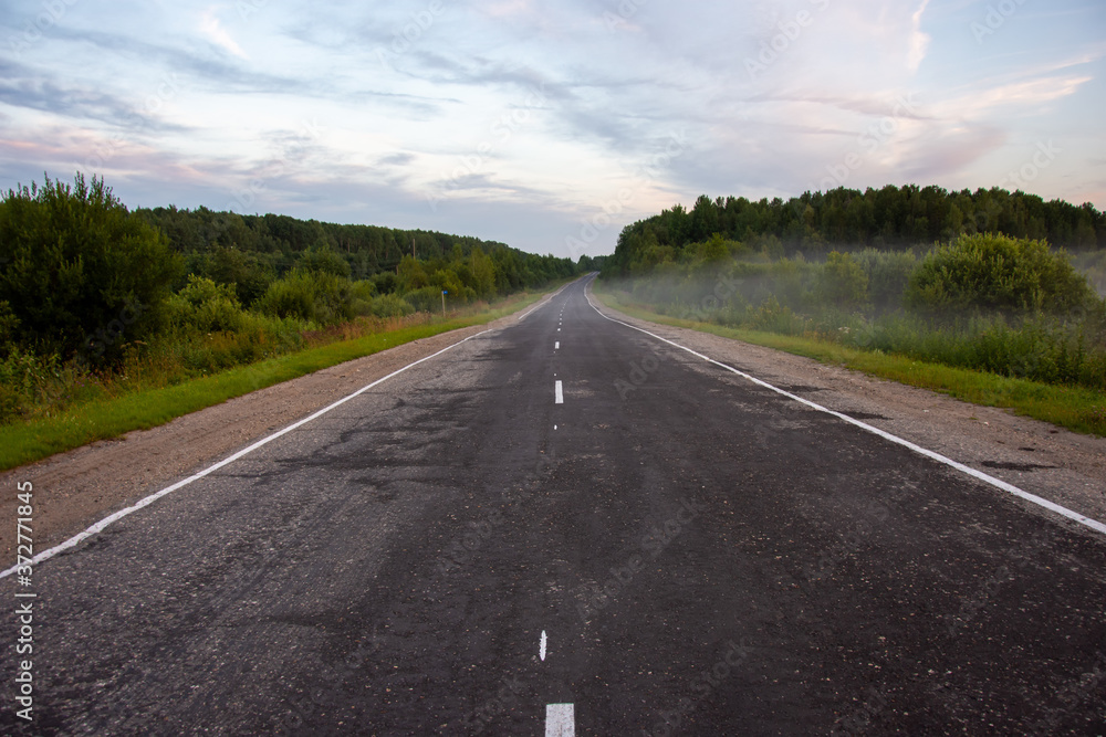 A view of the road going into the distance on a summer evening. Rural landscape with a road, trees, grass. There is fog in the background A view of the road going into the distance on a summer evening. Rural landscape with a road, trees, grass. There is fog in the background