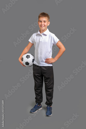 Kid holds in hand a classic soccer ball, looking at camera and smiling isolated on gray background. Full length, vertical orientation  © Serhii