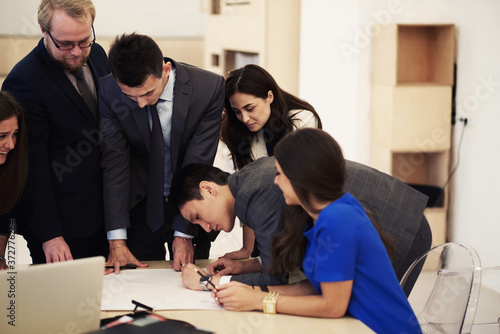 Group of a young creative architects reviewing blueprints during meeting in modern office interior, multi ethnic group of successful entrepreneurs in formal wear working together on joint project