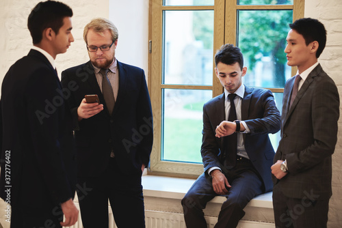 Group of young intelligent international students taking break between lectures in University, professional office workers resting after business meeting standing in corridor and waiting convocation