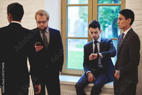 Group of young successful businessmen dressed in elegant suit having break during work day,confident financiers in luxury formal wear resting after conference while standing in office interior hallway