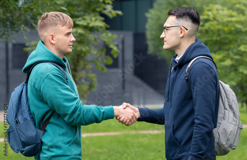 Two students, young men or guys shaking hands. University or college students, friends with backpacks greeting each other outdoors in park. Acquaintance, making a new friend. Handshaking.