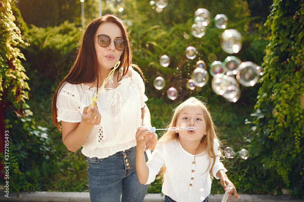 Family in a summer park. Mother in a blue pants. Little girl playing with bubbles.