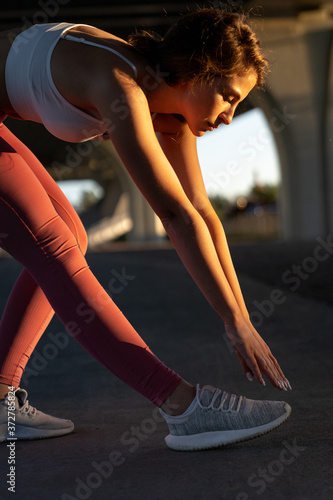 Slim sportswoman in pink leggings standing under the bridge at sunset, stretching muscles making functional training, makes a slope before a work out and jogging. Spine, hamstring stretch. 