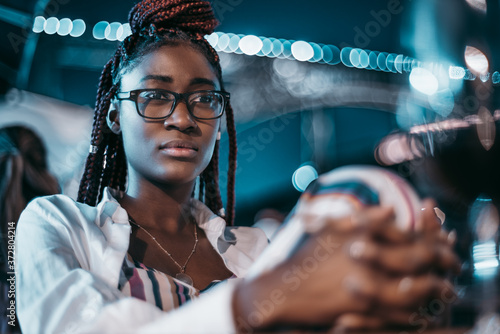 Night portrait of a beautiful young black woman in glasses sitting outdoors, holding her knee with hands and pensively looking into the distance with a silhouette of a bridge in a defocused background photo
