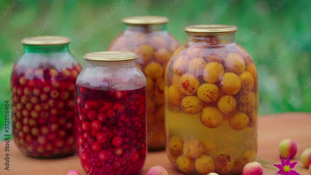 Close up of canned juice with fruits and berries on table in garden. Sealed jars of compote with apricots and flowers on table in yard. Concept of still life. Close up of canned juice with fruits and berries on table in garden. Sealed jars of compote with apricots and flowers on table in yard. Concept of still life.