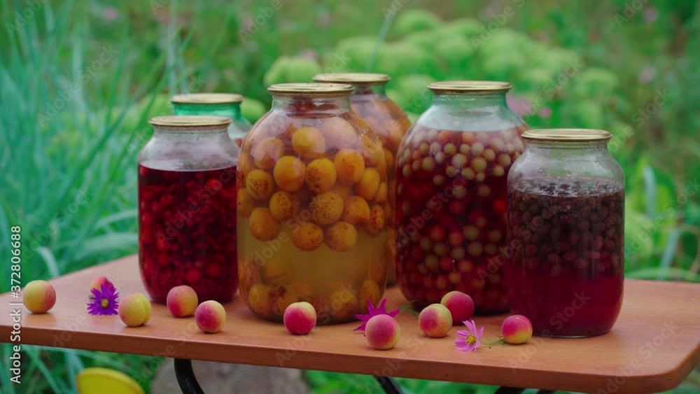 Close up of canned juice with fruits and berries on table in garden. Sealed jars of compote with apricots and flowers on table in yard. Concept of still life. Close up of canned juice with fruits and berries on table in garden. Sealed jars of compote with apricots and flowers on table in yard. Concept of still life.