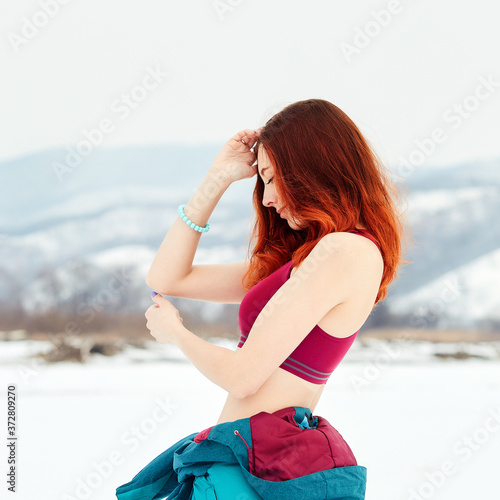 Gorgeous ginger women against the background of snow-covered river and mountains. Stylish sports young woman in a bright crimson top posing on a winter day